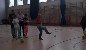 Osoby są na sali gimnastycznej. Mężczyzna stoi na jednej nodze a drugą ma w powietrzu. Za nim stoją ludzie.