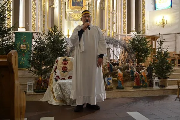 Mężczyzna stoi z mikrofonem w ręce. Jest ubrany w białą szatę.
