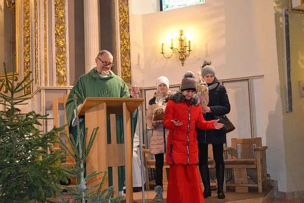 Ksiądz stoi na ambonie. Po prawej stronie stoją kobiety. Jedna z nich ma rozłożone ręce .