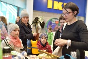 Kobiety i dzieci stoją przy stole na którym są ustawione różne artykuły. Kobieta po prawej stronie patrzy wprost na stół.