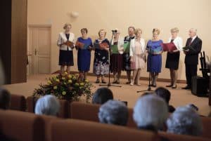 Na scenie jest grupa starszych osób. Wszyscy trzymają śpiewniki, a przed sobą mają mikrofon.Widownia jest tyłem do obiektywu.