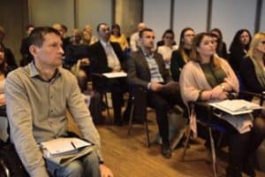 W sali siedzą osoby w rzędach. niektóre osoby mają na kolanach kartki i długopisy. Wszyscy patrzą przed siebie.