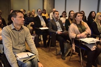 W sali siedzą osoby w rzędach. niektóre osoby mają na kolanach kartki i długopisy. Wszyscy patrzą przed siebie.