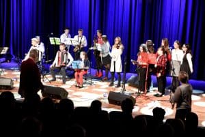 Na scenie stoi grupa osób. Po prawej stronie są to dziewczyny stojące przed śpiewnikiem. Po lewej stronie siedzi dwóch chłopców z instrumentami. Za nimi stoją chłopcy i dziewczęta z instrumentami.