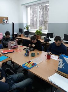 Grupa mężczyzn siedzi przy dużym stole. Trzech z nich jest pochylonych i patrzą na blat. Na stole są kolorowe kartki, długopisy. Jeden z nich jest przycięty w kadrze.