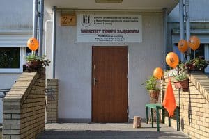 Wejście do budynku Warsztatów Terapii Zajęciowej. Po lewej i prawej stronie są pomarańczowe balony. Są włożone w doniczki z roślinami.