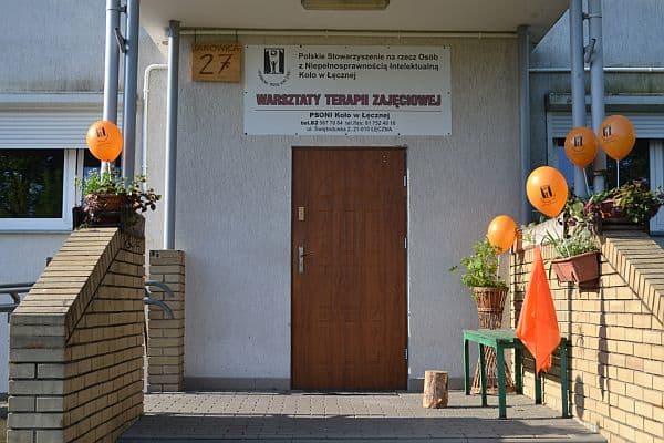 Wejście do budynku Warsztatów Terapii Zajęciowej. Po lewej i prawej stronie są pomarańczowe balony. Są włożone w doniczki z roślinami.