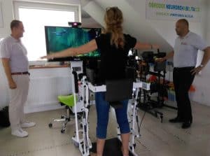 Kobieta stoi na urządzeniu rehabilitacyjnym. Ręce trzyma wyciągnięte na boki i patrzy w ekran. Po jej obu stronach stoją mężczyźni.