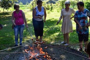 Kobiety stoją przy palenisku. Trzymają długie patyki na których nabite są kiełbaski i smażą je.