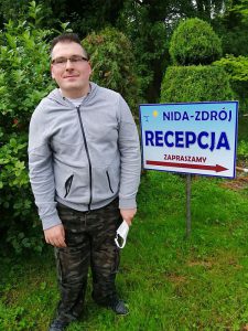 Mężczyzna pozuje do zdjęcia na trawniku. Obok tabliczka z napisem: “Nida- Zdrój. Recepcja. Zapraszamy”.