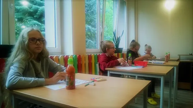 Czworo dzieci siedzi w ławce. Przed soba maja biała kartkę i soki. Dziewczynka najbardziej z przodu patrzy się w obiektyw.