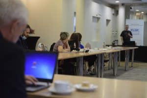 Po lewej stronie siedzi mężczyzna tyłem do obiektywu i ma otwarty laptop. Przy stole po lewej stronie siedzą ludzie. Na końcu stoi mężczyzna z mikrofonem.