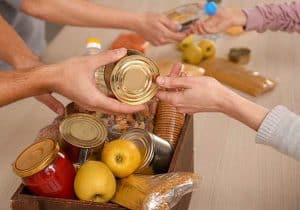 Na stole w drewnianej skrzyneczce leżą różne produkty żywnościowe: jabłka, słoiki, ciastka. Nad nimi są ręce osób, które przekazują sobie produkty. Obok na stole leżą jabłka i inne produkty.