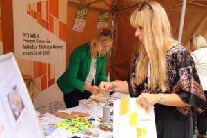 Kobieta stoi przed stołem. Trzyma torbę biało-żółta. Za stołem stoi kobieta, która pochyla się. Za nia jest baner z napisem: Power Program Operacyjny Wiedza Edukacja Rozwój na lata 2014-2020.
