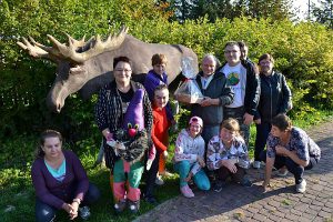 Grupa osób pozuje do zdjęcia przy figurze łosia. Starszy mężczyzna trzyma zapakowane pudełko.