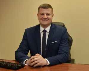 Mężczyzna w garniturze siedzi przy biurku i uśmiecha się do zdjęcia. Na biurku klawiatura komputerowa