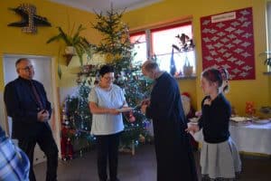 Kobieta trzyma opłatek. Obok niej stoi ksiądz w maseczce. Po prawej stronie za księdzem stoi dziewczynka, a po lewej za kobietą mężczyzna. W tle choinka