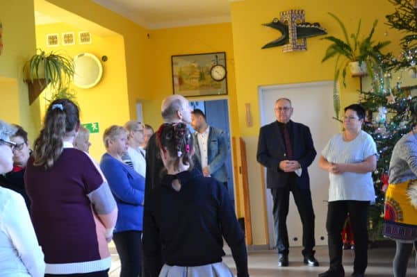 Grupa osób stoi w sali. Przy choince po prawej stronie stoi kobieta i mężczyzna. Po lewej widać dzieci, które są odwrócone do obiektywu tyłem.