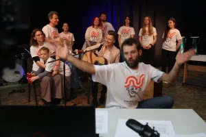 Grupa osób stoi. Przed mikrofonem siedzi kobieta z gitarą, a obok kobieta siedzi i trzyma pochylające się dziecko. Mężczyzna najbardziej z przodu siedzi przed fortepianem i trzyma ręce w górze.