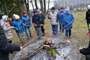 Uczestnicy stoją w półkolu i przypiekają nad ogniskiem kiełbaski nadziane na patyku.