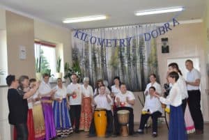 18-osobowy zespół klaszcze rytm wykonywanej piosenki. Kobiety są ubrane w białe bluzki i różnokolorowe długie spódnice, a mężczyźni w białe koszulki i ciemne spodnie. Nad nimi wywieszony jest napis Kilometry Dobra.