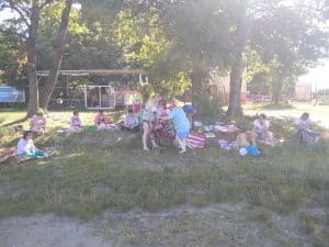 Liczna grupa uczestników odpoczywających na trawie na rozłożonych kocach w cieniu drzew. Na pierwszym planie widać dwie stojące kobiety, z czego jedna z balkonikiem.