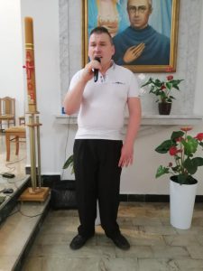 Młody mężczyzna śpiewa do mikrofonu. W tle boczny ołtarz z połową obrazu przedstawiającego świętego zakonnika w okularach i tułów osoby w niebieskim stroju trzymającą w ręce koronę. Obok obrazu świeca paschalna.