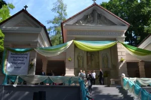 Przy kaplicy Matki Bożej stoją ludzie. Budynek jest starodawny i przyozdobiony niebiesko- biało- żółtym materiałem.