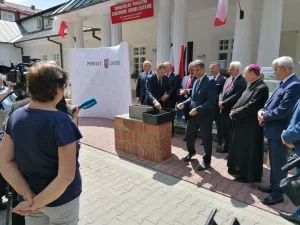 Grupa mężczyzn stoi przed budynkiem. Jeden z nich nakłada zaprawę do pustaka. Przed nimi stoją dziennikarze.