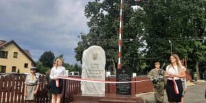 Dwie młode kobiety stoją przed pomnikiem i trzymają biało-czerwoną wstęgę. Za nimi stoi dwóch mężczyzn ubranych w wojskowe mundury polowe. Za kobietą po lewej stronie stoi harcerka.