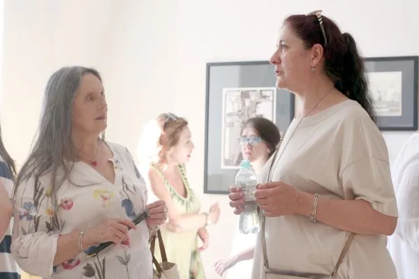 Dwie panie w średnim wieku rozmawiają. W tle inne kobiety podziwiające obrazy na ścianie.