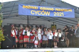 Uczestnicy stoją na scenie i pokazują zdobyte dyplomy. Wśród nich są dzieci oraz kobiety ubrane w stroje ludowe.