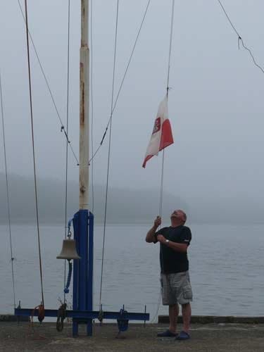 Na zdjęciu mężczyzna w niebeiskiej koszulce i białych spodenkach wciąga na maszt flagę. Mglisty dzień.