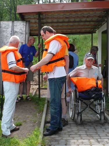 Na zdjęciu trzej mężczyźni w tym jeden na wózku zakładają kamizelki ratunkowe.