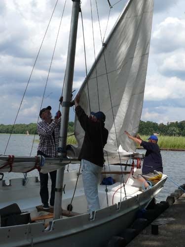 Na zdjęciu przygotowania do wypłynięcia na wody jeziora. Dwaj mężczyźni wciągają na maszt żagle.