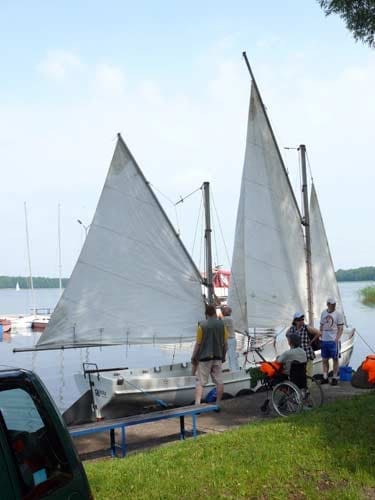 Na zdjęciu gotowa żaglówka do rejsu. Na nabrzeżu grupa osób w tym jedna na wózku oczekuje wejścia na pokład.
