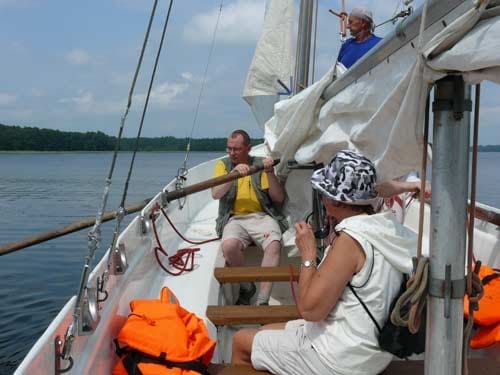 Na zdjęciu mężczyzna wiosłuje, naprzeciw niego siedzi kobieta. W tle mężczyzna majstruje przy linkach masztu.