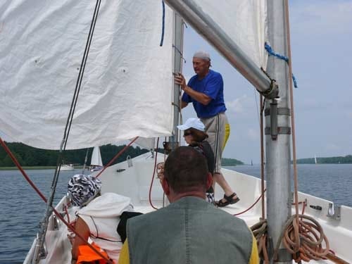 Na zdjęciu plecami do fotografa mężczyzna przygląda się drugiemu jak ten stawia żagiel na maszt.