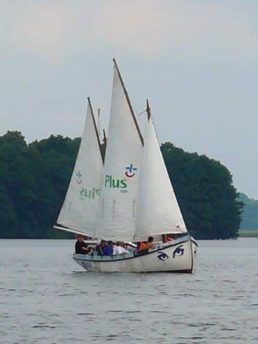 Na zdjęciu płynąca żaglówka z pełnymi żaglami z logiem Plusa.