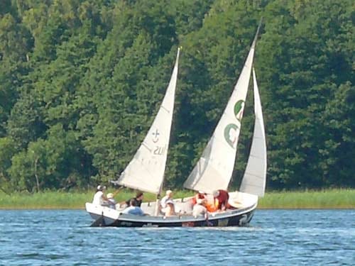 Na zdjęciu płynąca żaglówka z pełnymi żaglami z logiem Plusa.