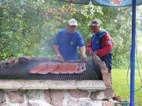 Na zdjęciu dwaj mężczyźni w czapkach z daszkiem dogląda dużej ilości kiełbasek na dużym grillu.