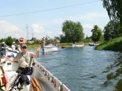 Na zdjęciu Kazimierz Adomat w okularach przeciwsłonecznych i w kaszkietówce siedzi na burcie i macha do fotografa. U jego nóg stoi złożony wózek inwalidzki. W tle wody przystani.