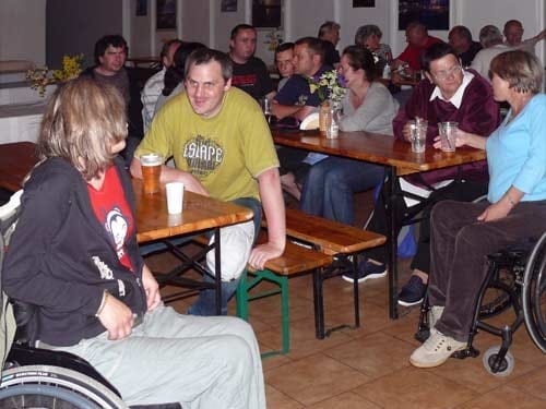 Na zdjęciu grupa ludzi siedzi na ławach przy stołach. Pija piwo i rozmawiają między sobą. Po lewej stronie zdjęcia mężczyzna rozmawia z kobietą na wózku.