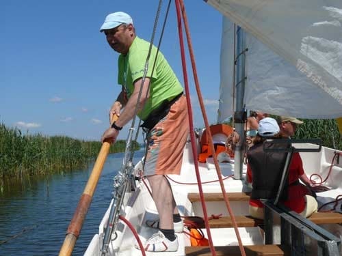 Na zdjęciu mężczyzna w zielonej koszulce, pomarańczowych szortach i w niebiskiej czapce z daszkiem odpycha wiosłem od dna by ruszyć łódkę.