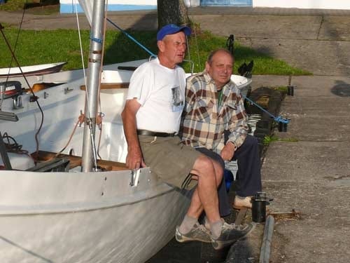 Na zdjęciu dwaj mężczyźni siedzą na burcie łodzi na brzegu i obejmują się przyjaźnie.