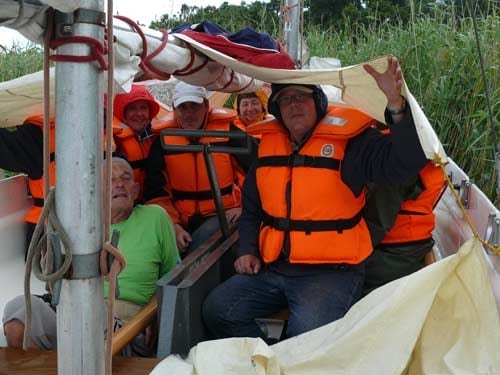 Na zdjęciu grupa uczestnikó w pomarańczwych kamizelkach chroni się przed deszczem przykrywajac sie pod dużą płachtą - żaglem