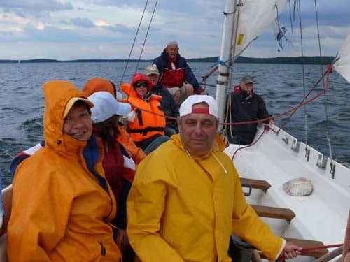 Na zdjęciu grupa osób w żółtych kurtkach przeciwdeszczowych płynie żaglówką.