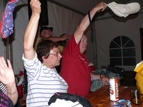 Na zdjęciu kobieta w białej bluzce i w okularach oraz mężczyzna w czerwonej koszulce wywijają czapkami nad głową. Entyzjastycznie odbierają grana muzykę.