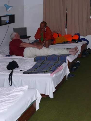 Na zdjęciu dwaj mężczyźni rozmawiają ze sobą siedząc lub leżąc na łóżkach.