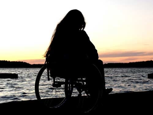 Na zdjęciu czarna sylwetka kobiety na wózku, która podziwia zachód słońca nad wodą.